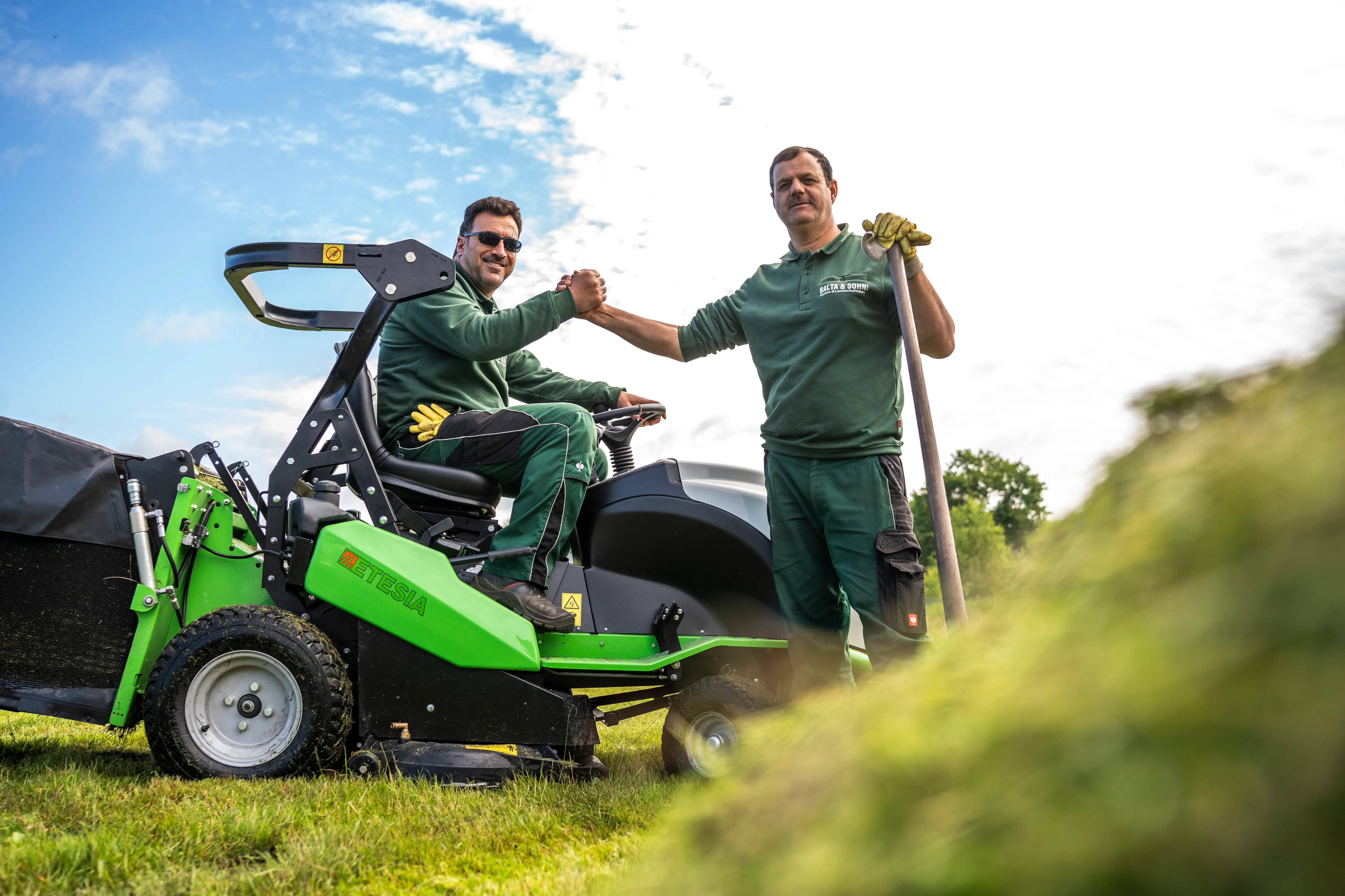 Zwei Gärtner in grüner Arbeitskleidung geben sich die Hand vor einem grünen Aufsitz-Rasenmäher im Freien.