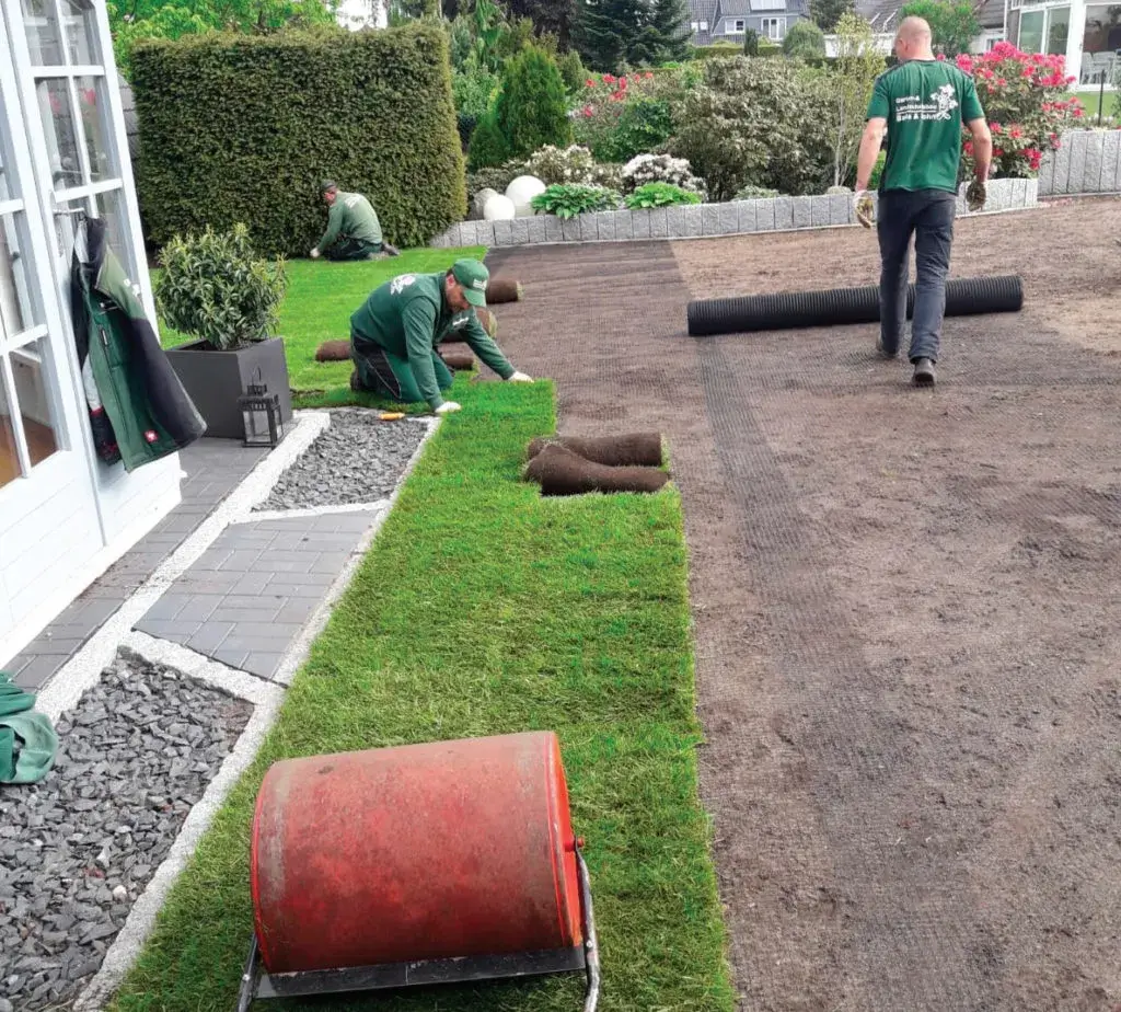 Gartenarbeiter verlegen Rollrasen auf einem Grundstück neben einem Haus mit gepflastertem Weg.
