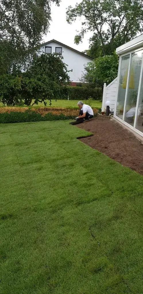 Person verlegt Rollrasen an einem Haus mit Wintergarten im Garten.