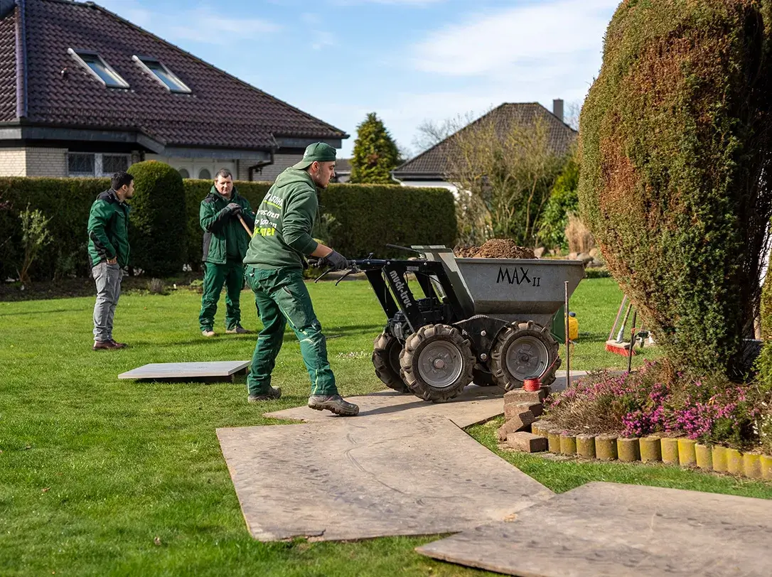 Gärtner schieben eine kleine Kipplastmaschine mit Erde auf einem Rasenweg, im Hintergrund stehen zwei weitere Männer vor einem Haus.