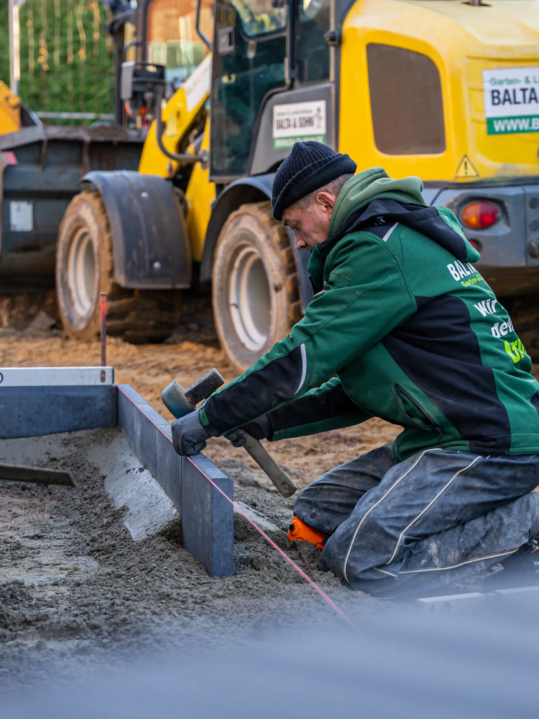 Arbeiter kniet auf Boden und richtet Pflastersteine mit Hammer und Wasserwaage aus, Bagger im Hintergrund.