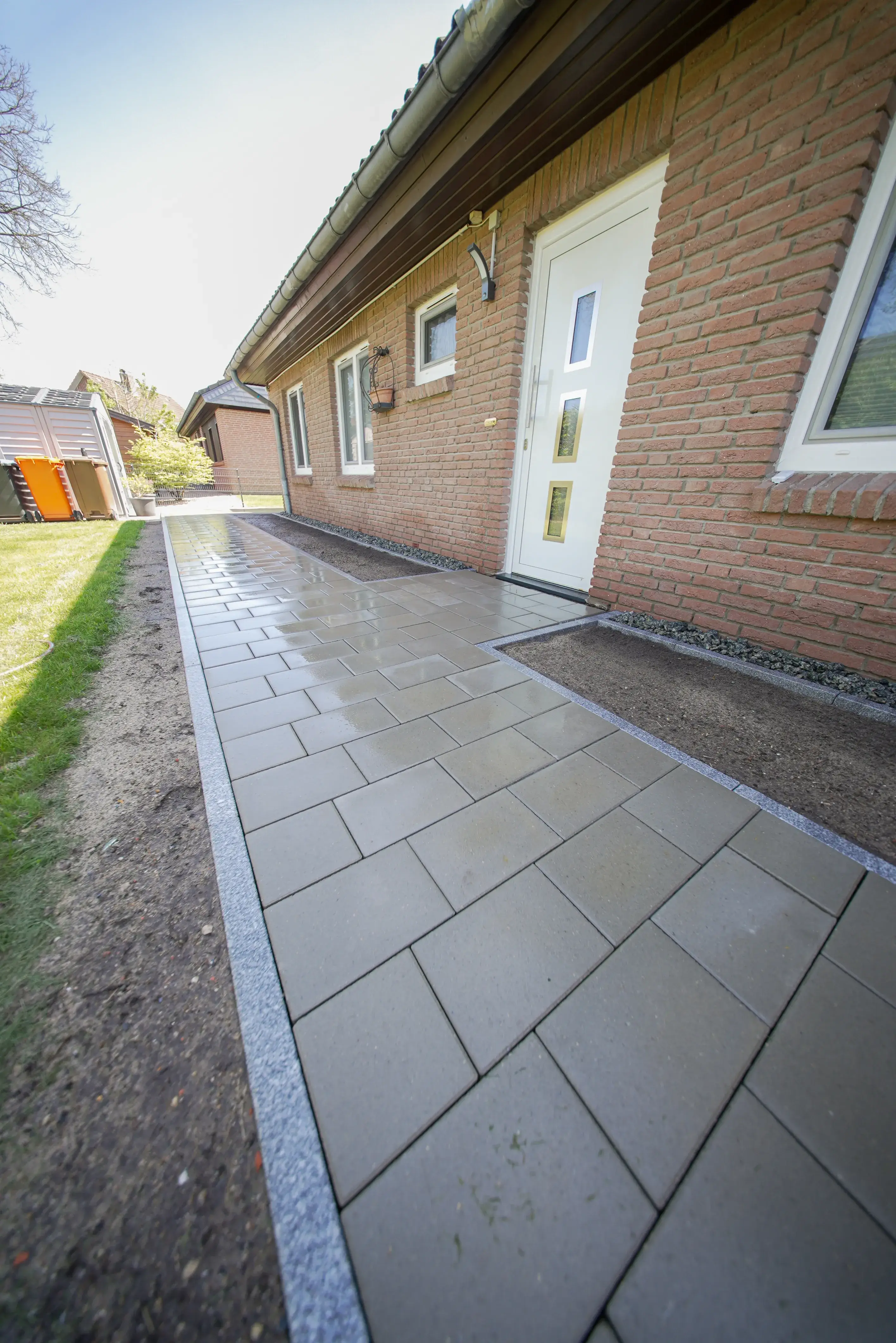 Nasser, neu verlegter Gehweg aus rechteckigen grauen Pflastersteinen entlang eines Backsteinhauses.