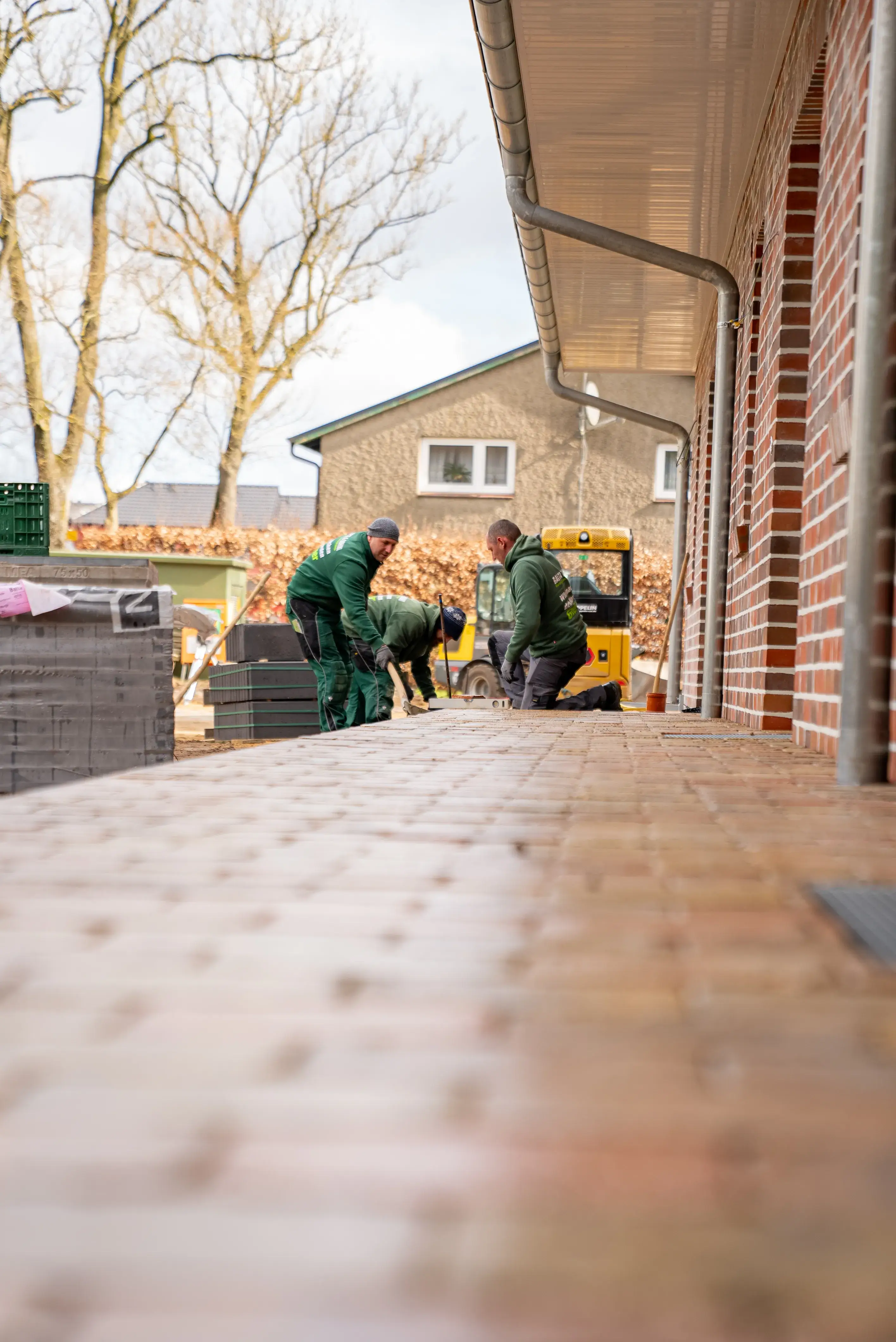 Drei Bauarbeiter verlegen Pflastersteine vor einem roten Backsteingebäude an einem sonnigen Tag.