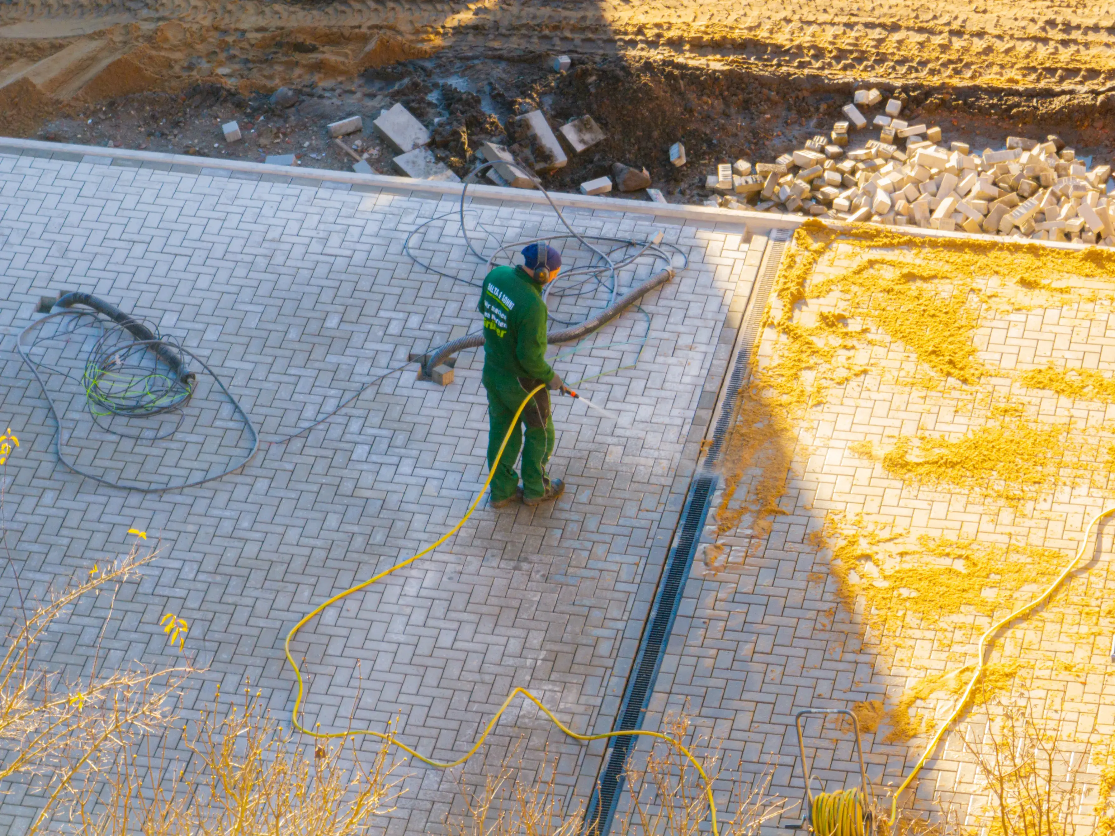 Ein Arbeiter reinigt eine gepflasterte Fläche mit einem Hochdruckreiniger, neben Sand und gestapelten Pflastersteinen.