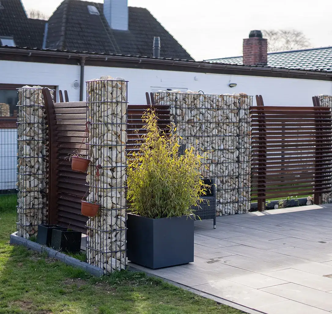 Moderner Gartenzaun aus Holzlatten und Steinsäulen mit Eisendraht, davor ein großer Pflanzkübel mit grünem Busch.