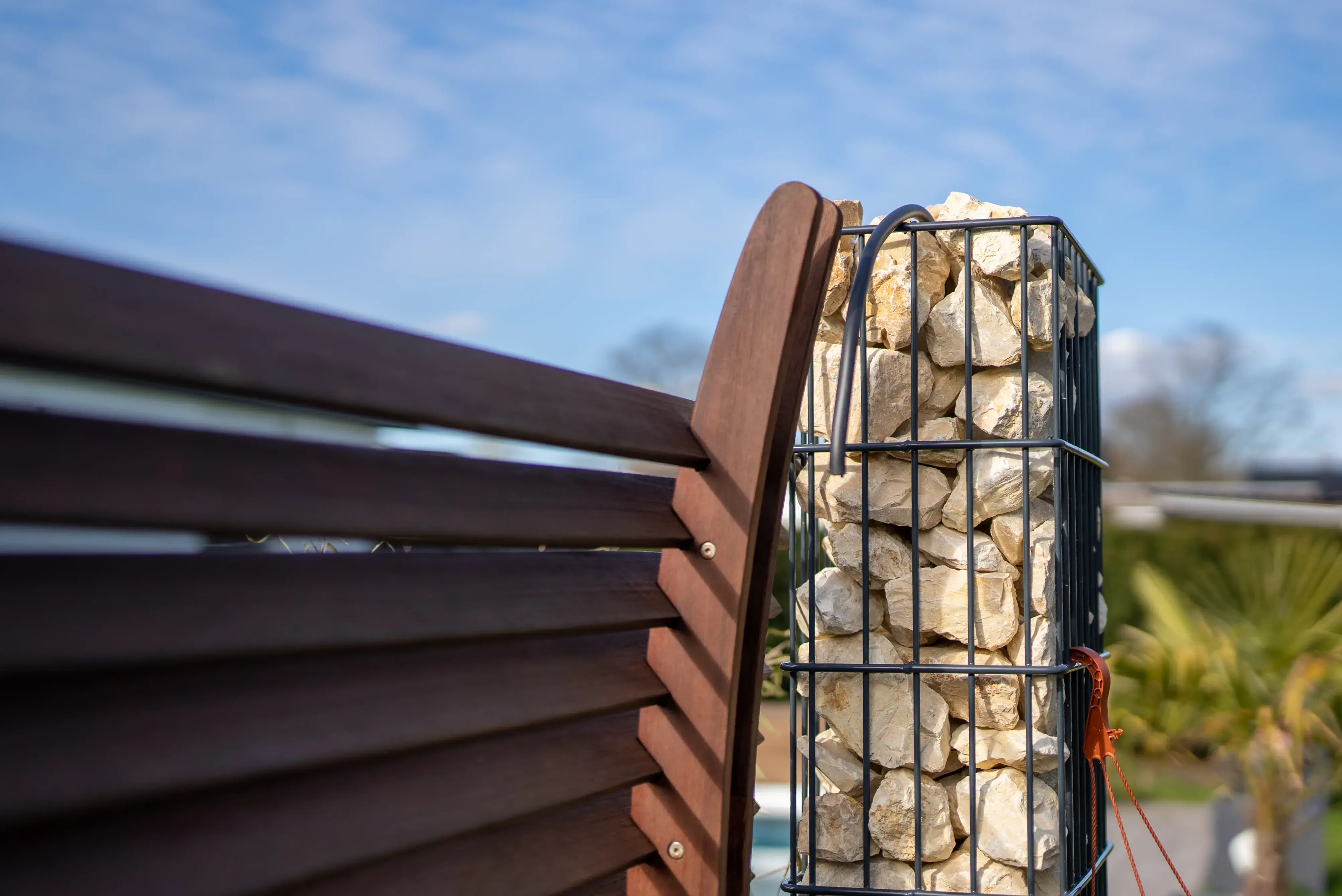 Nahaufnahme eines Holzzauns neben einer Säule aus Steinen in einem Metallgitter bei sonnigem Himmel.