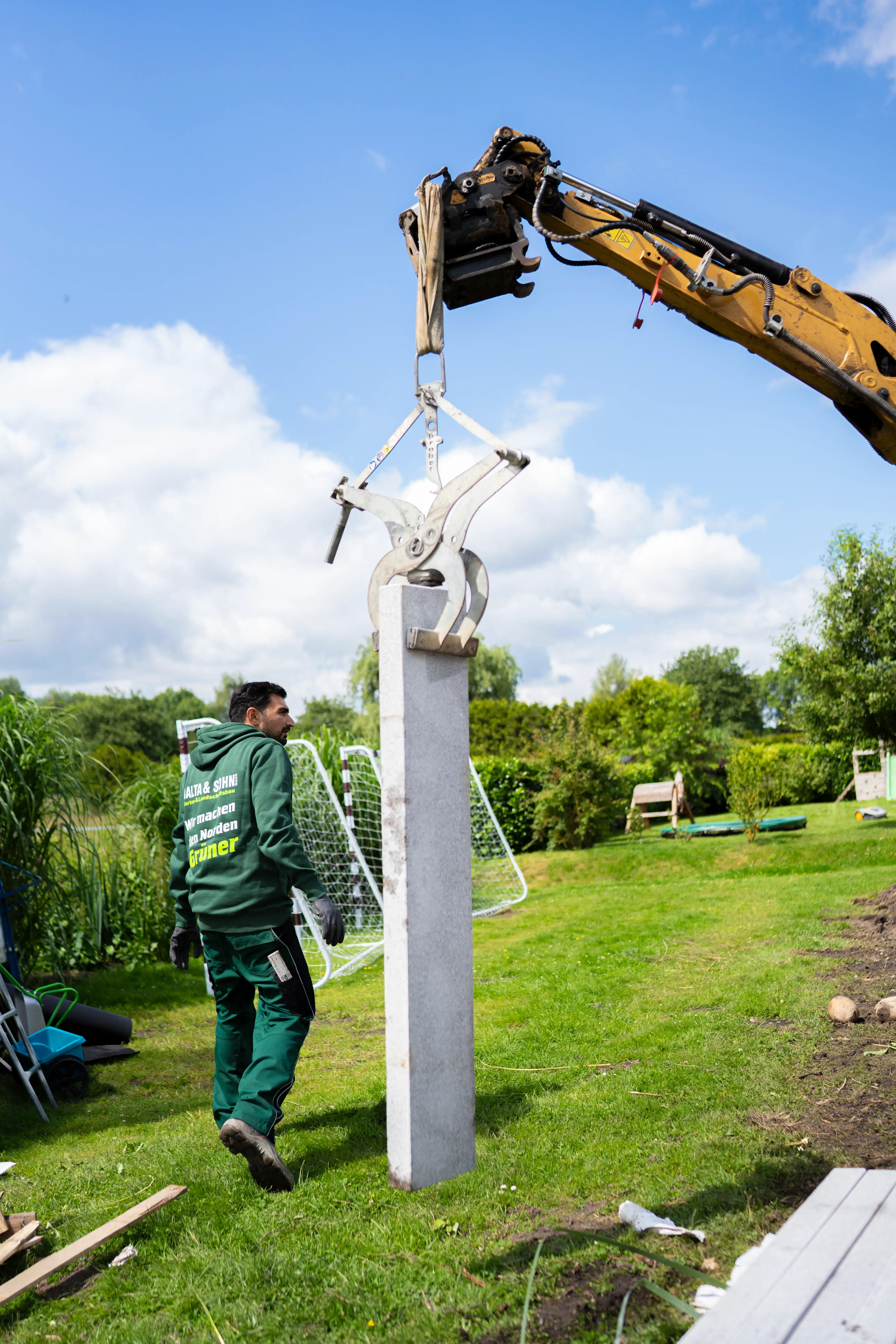 Gärtner trägt grüne Arbeitskleidung und überwacht Hebevorrichtung, die großen grauen Zaunpfahl an Baggerarm hält.