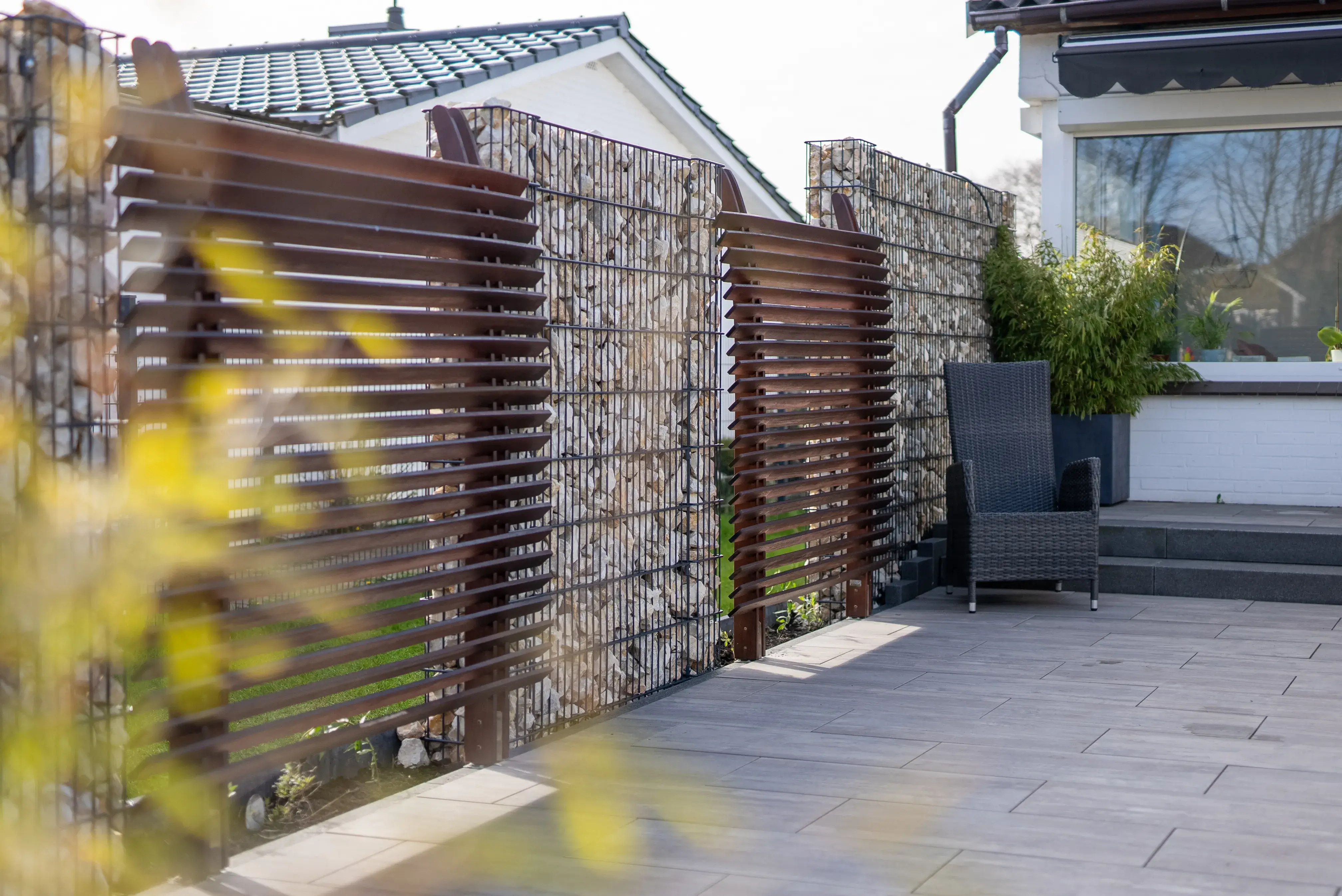 Moderner Gartenzaun mit Gabionen aus Steinen und braunen Holzlatten neben einer Terrasse mit grauem Sessel.