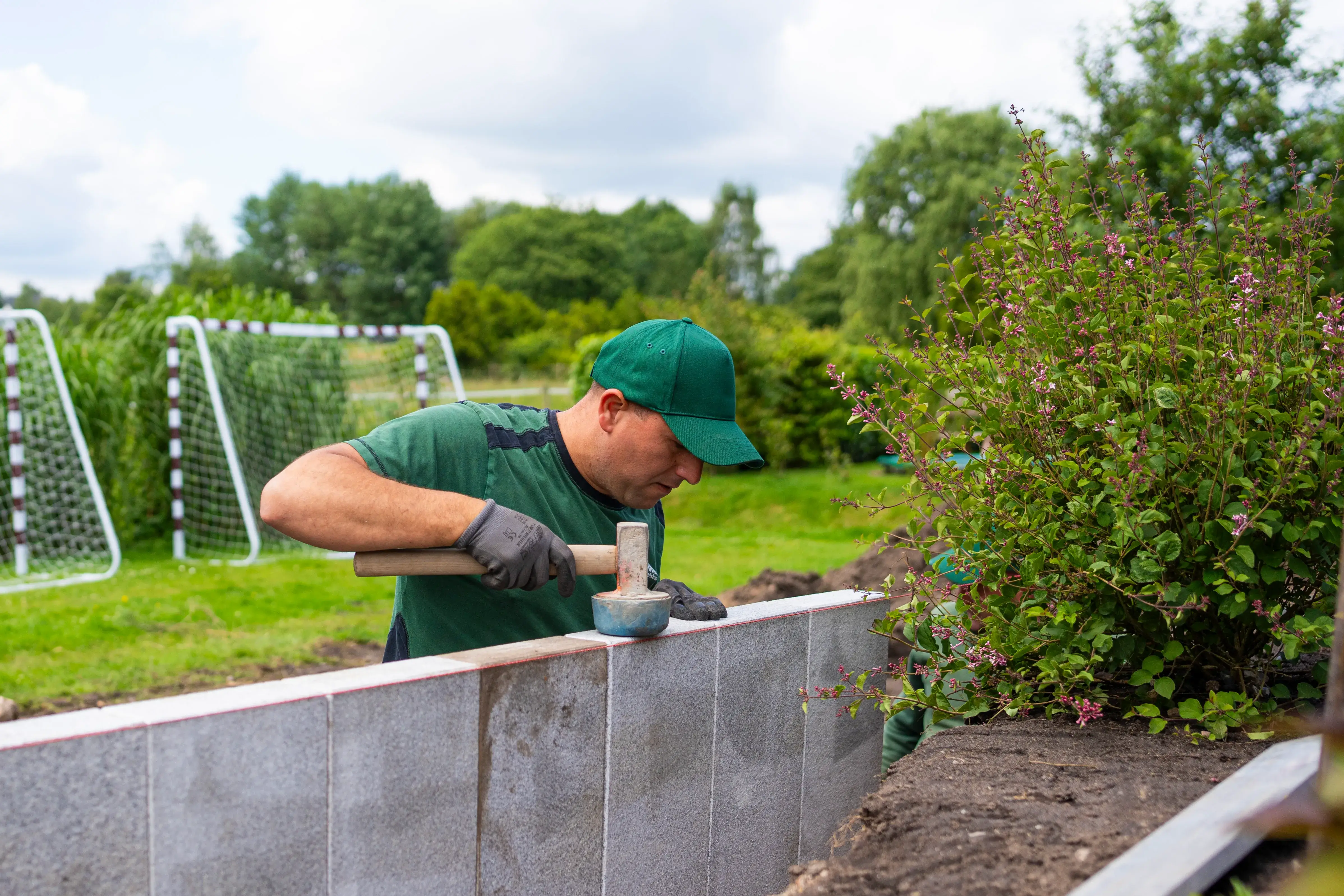 Mann trägt grüne Kappe und arbeitet mit Hammer und Gummihammer an einer grauen Betonmauer im Garten.