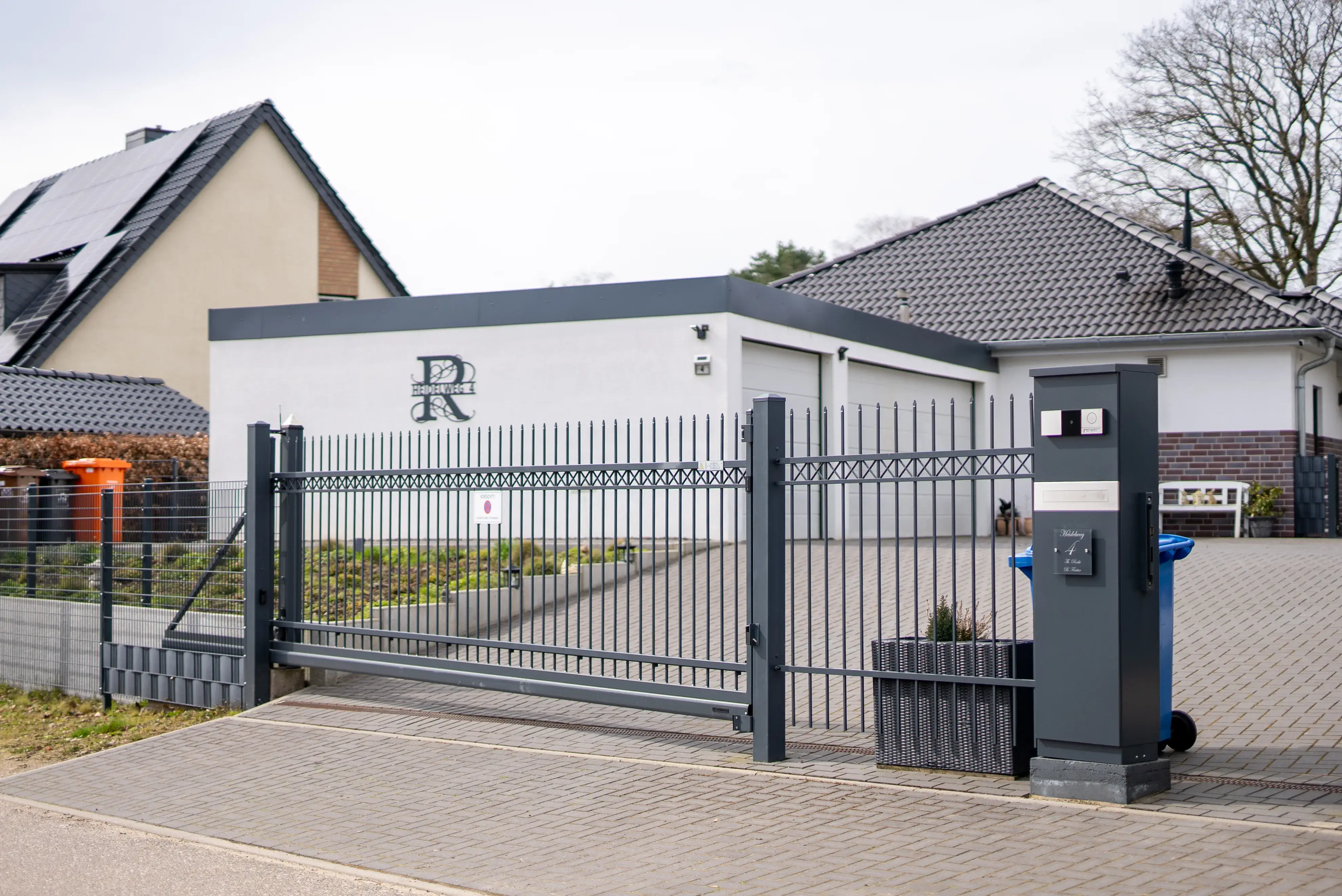 Moderner Einfahrtstor aus Metall mit automatischem Öffnungsmechanismus vor einem Einfamilienhaus mit Pflasterauffahrt.