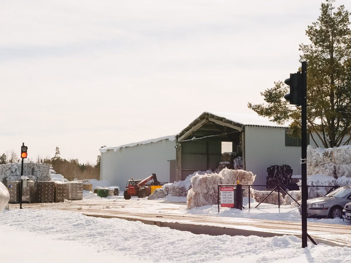 Snow-covered industrial yard with packed materials, a small red loader, and a large white storage shed under cloudy sky.
