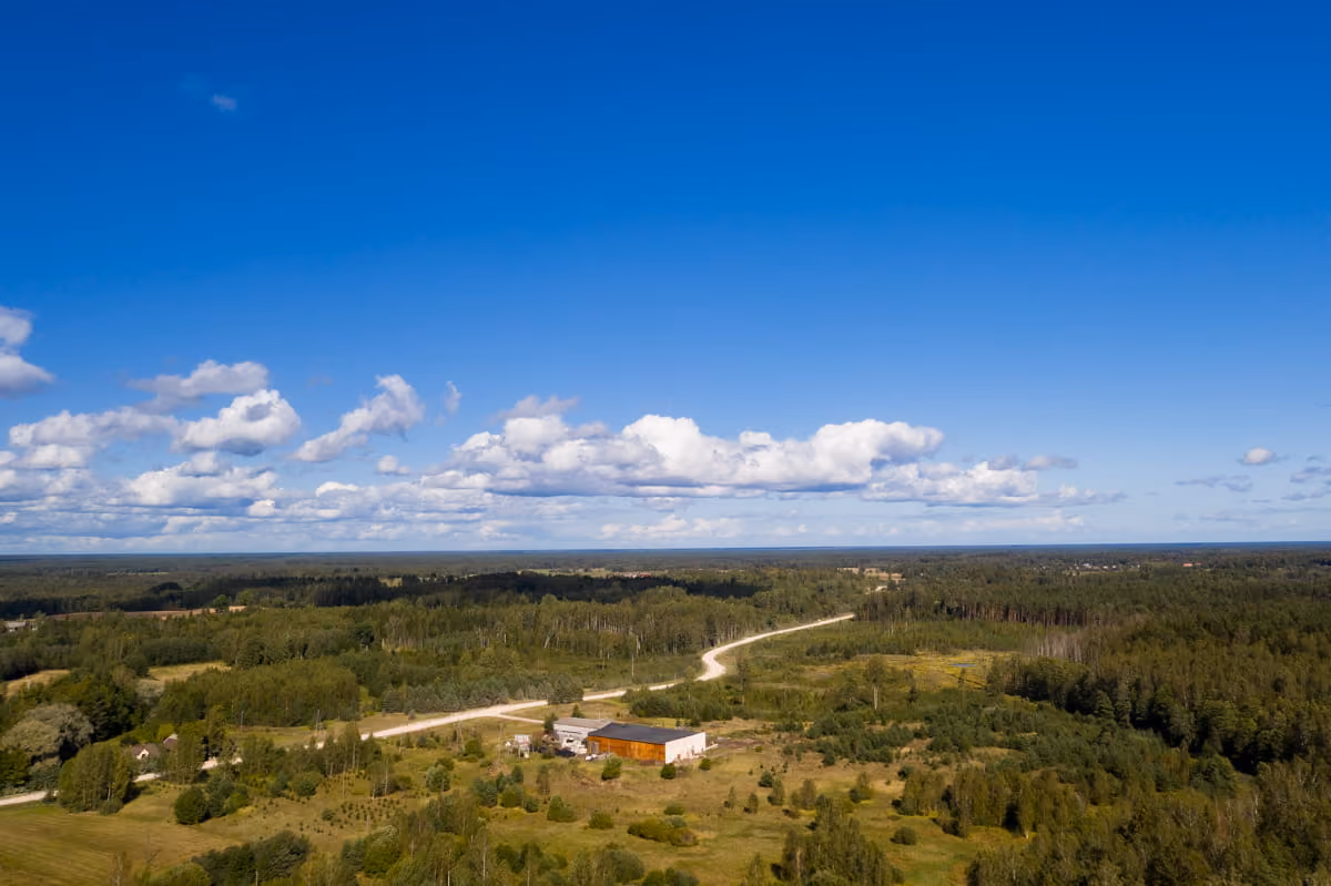 Aerial view of rural landscape with a winding dirt road, scattered buildings, and dense forest under a bright blue sky with scattered clouds.
