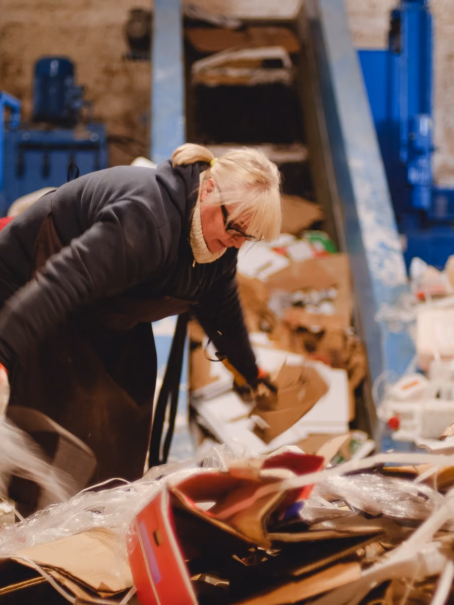 Woman wearing glasses and a dark jacket sorting cardboard and plastic waste in a recycling facility.