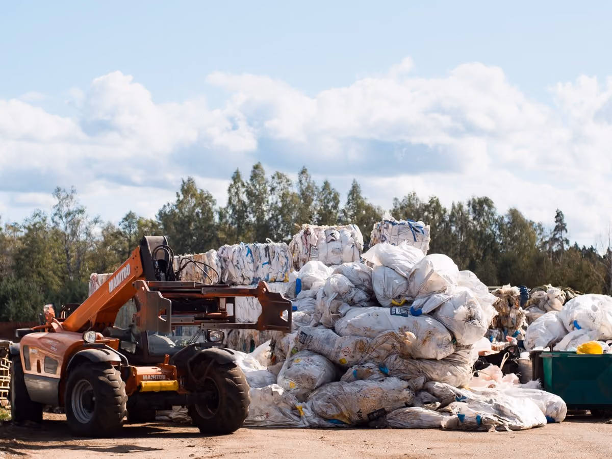 Orange forklift at a waste recycling site with large piles of compressed white plastic bags and waste materials stacked outdoors.