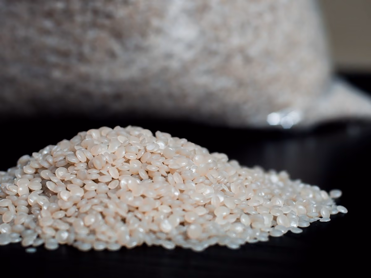 Close-up of a pile of uncooked polished white rice grains on a dark surface with a blurred bag of rice in the background.