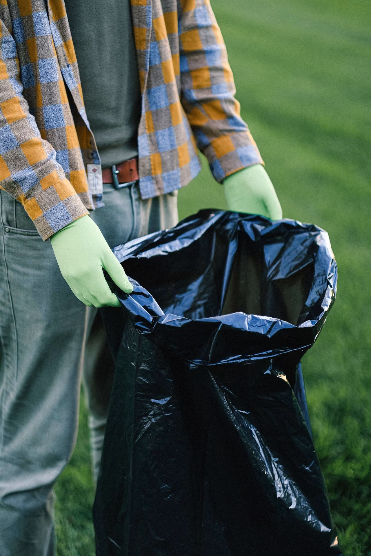 Person wearing green gloves holding open a large black trash bag outdoors on grass.