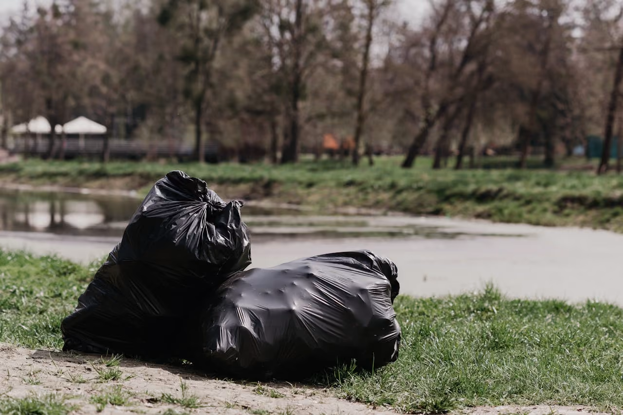 Two black garbage bags placed on grass near a body of water with trees in the background.