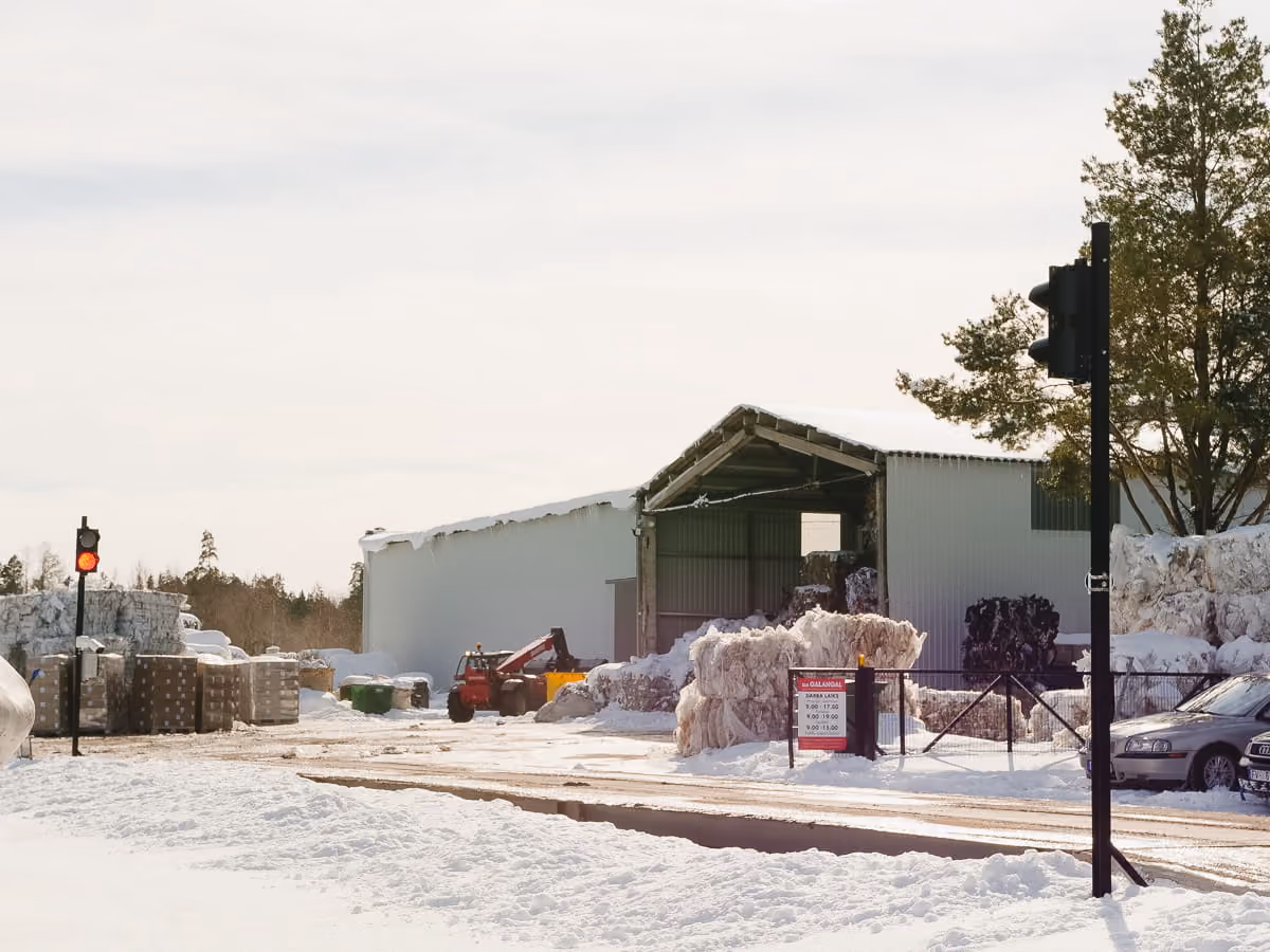 Snow-covered industrial yard with large stacked materials, a warehouse, and a red forklift under overcast sky.