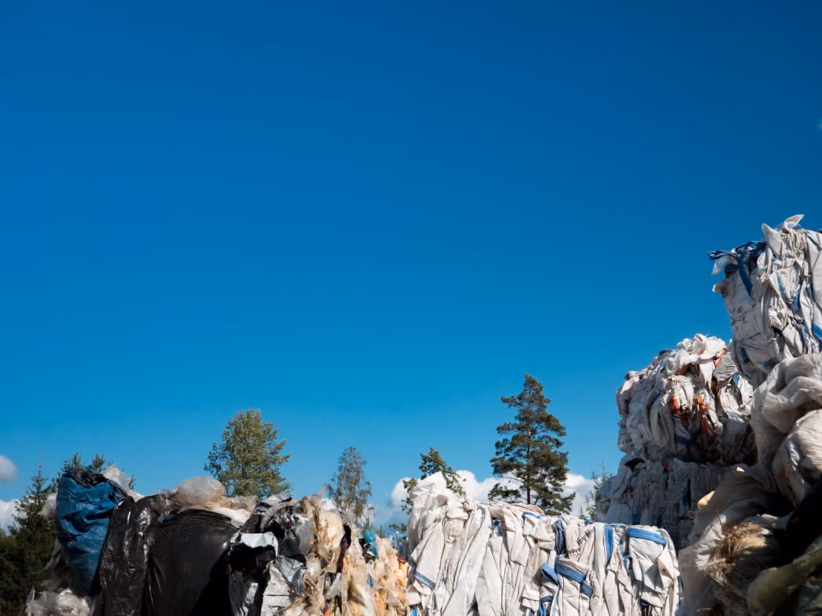 Large stacks of compressed plastic waste bales outdoors under a clear blue sky with trees in the background.