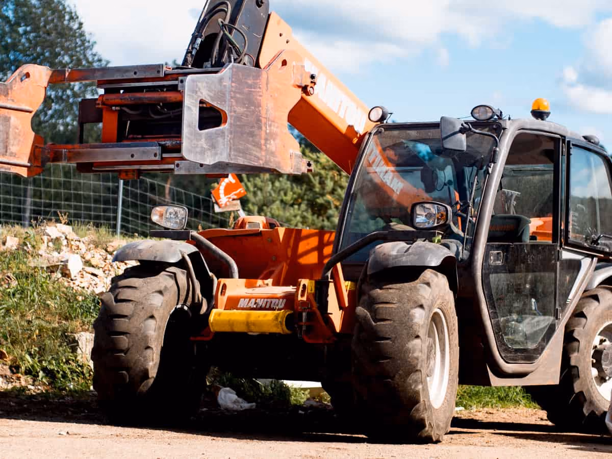 Orange Manitou telescopic handler with large tires and extended forks parked on a dirt path.