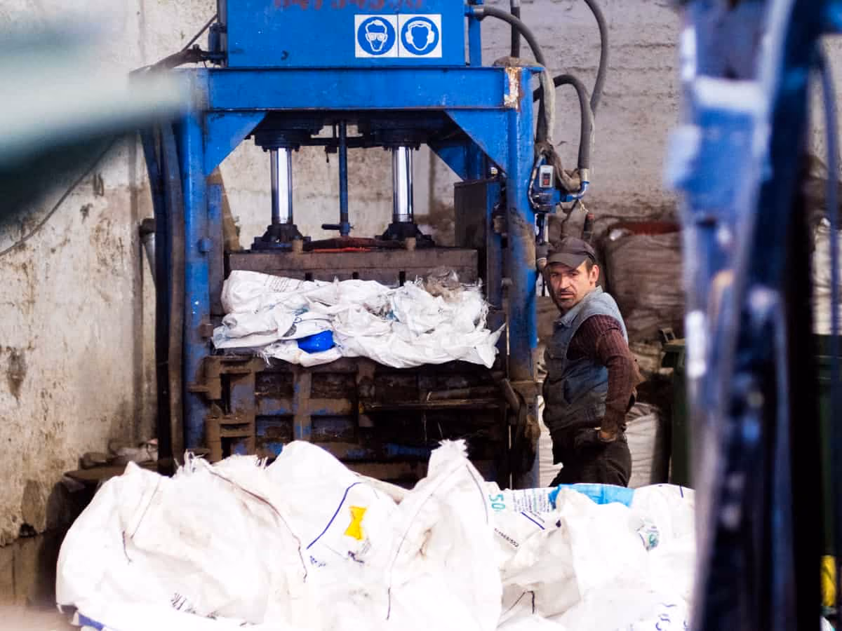 Worker standing next to a blue industrial machine with large white bags in a factory setting.