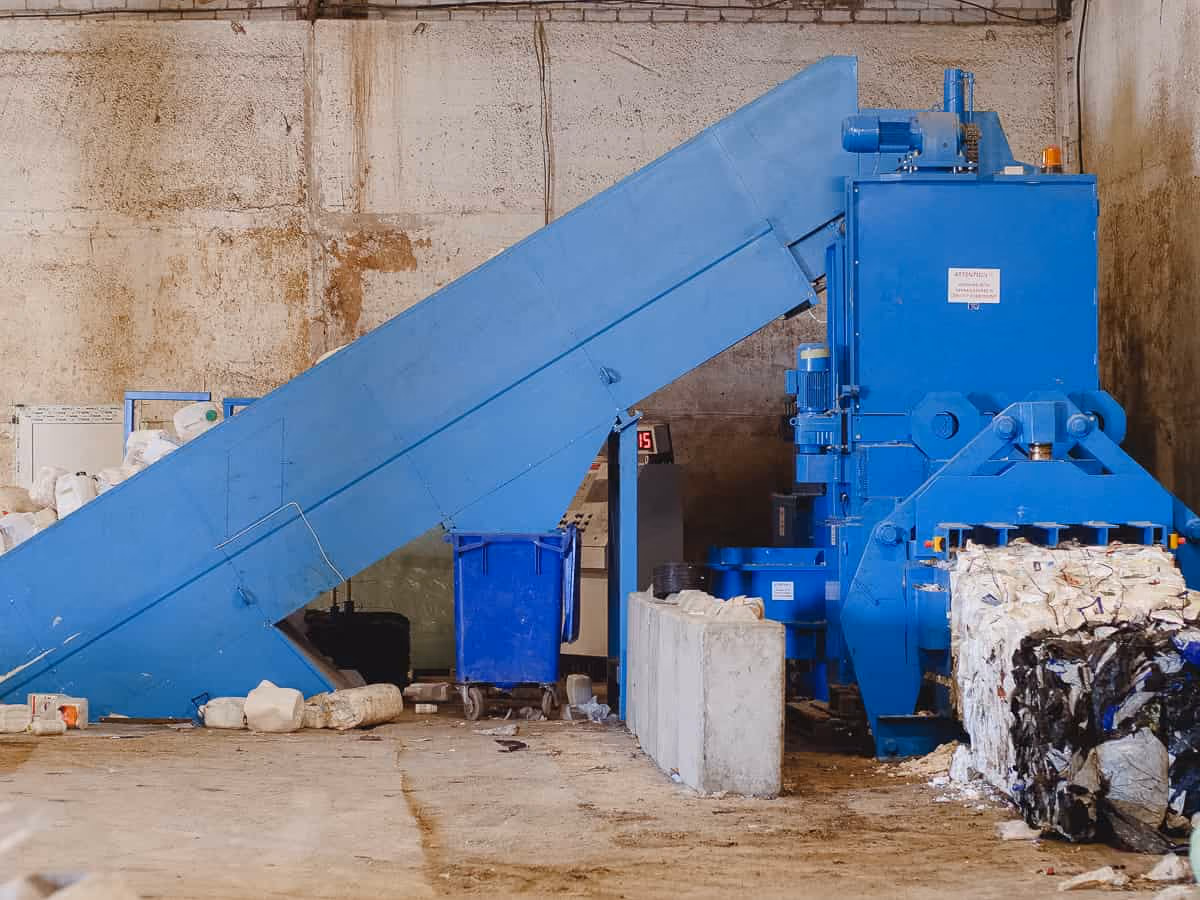 Blue industrial machine compressing and baling plastic waste inside a warehouse.