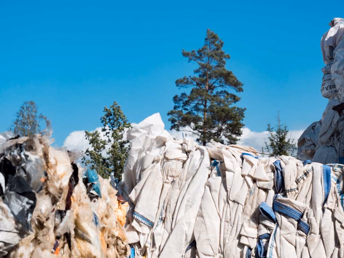 Large bales of compressed white and blue fabric stacked outdoors under a clear blue sky with trees in the background.