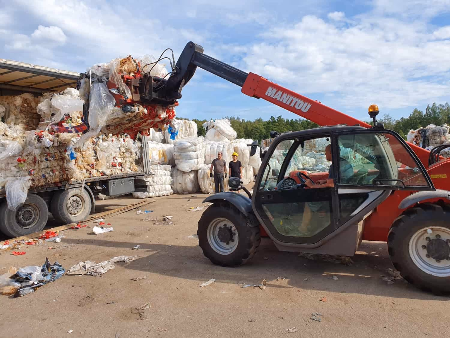Red Manitou telehandler lifting compressed plastic waste bales into a truck with two workers standing nearby.