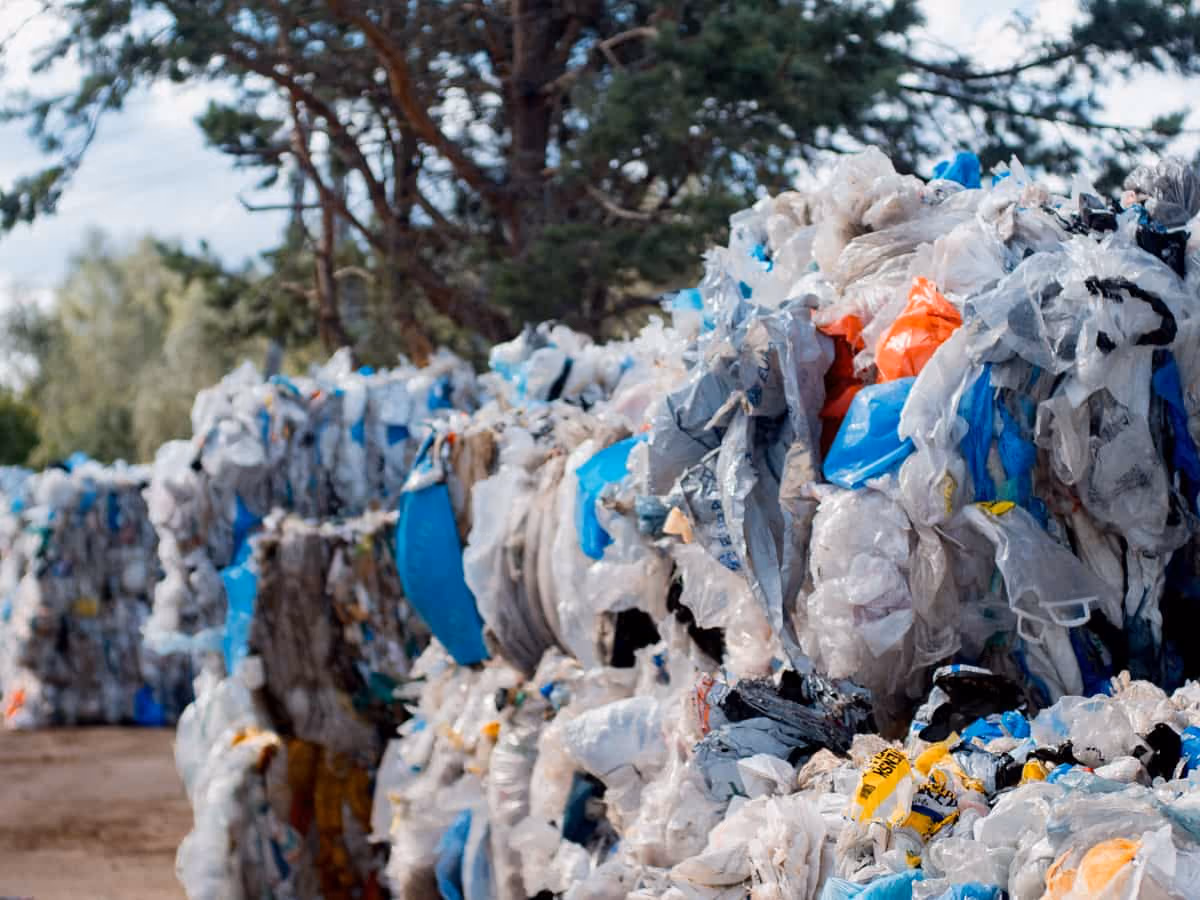 Stacks of compressed plastic waste bales outdoors with trees in the background.