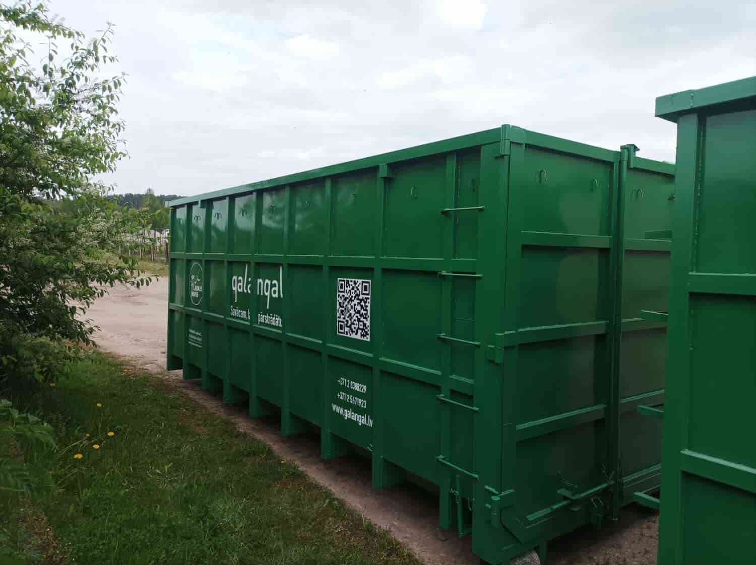 Large green metal waste container with company branding and a QR code on an outdoor path beside grass and trees.