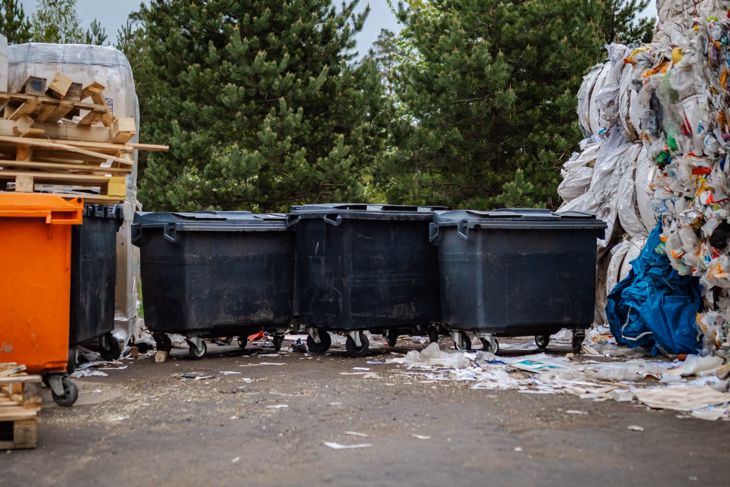 Several large black and one orange garbage containers outside near stacked wooden pallets and shredded plastic waste.