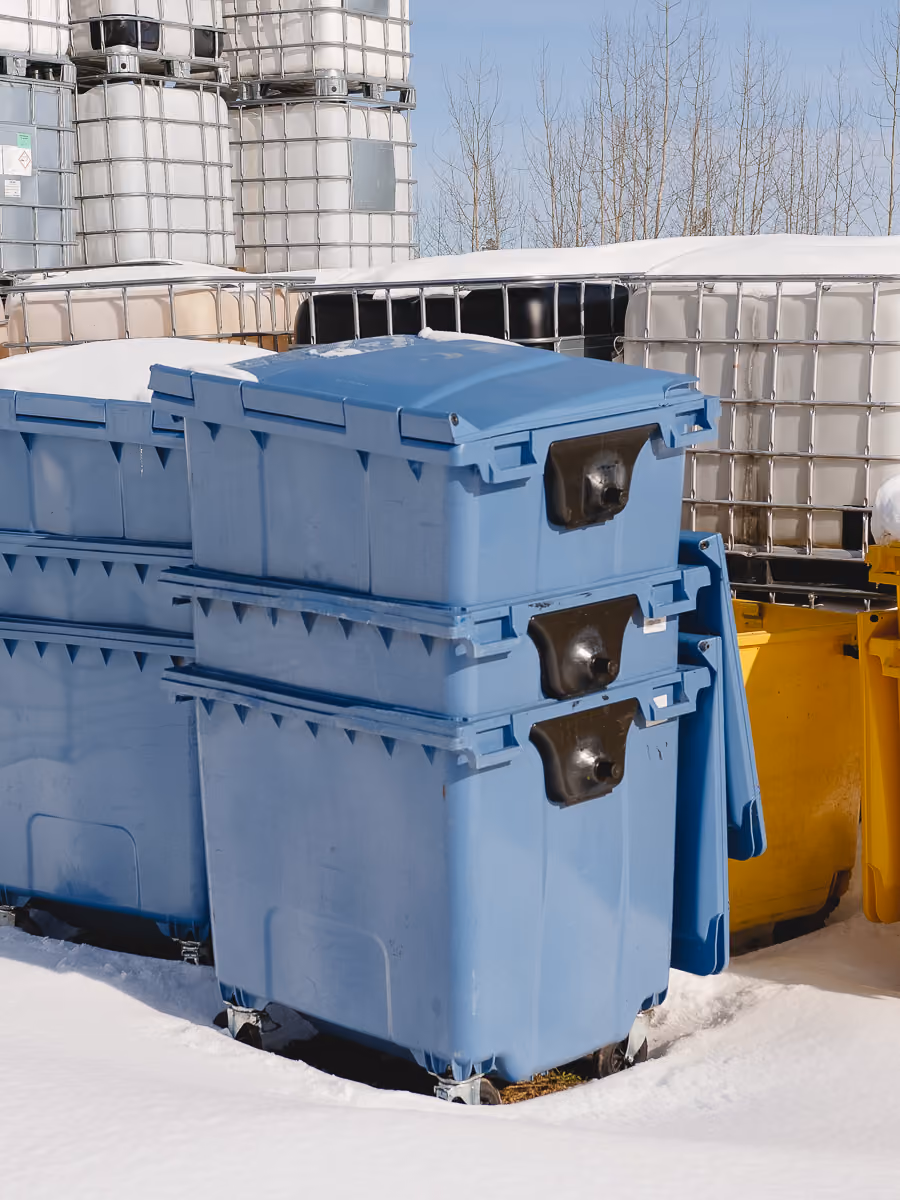 Stacked blue plastic waste bins on snow-covered ground with large white containers in the background.
