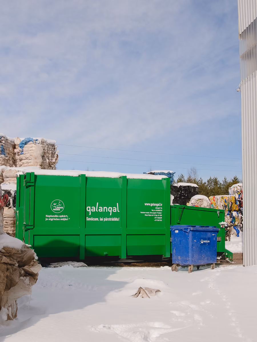 Green and blue recycling containers with snow and piled compressed recyclables outdoors.
