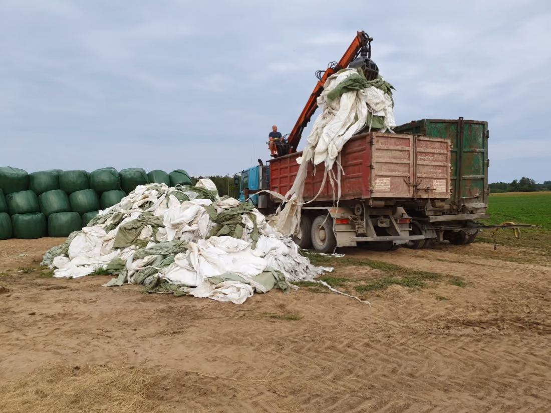 Farm truck unloading large white and green tarps with a man operating crane on a dirt field.