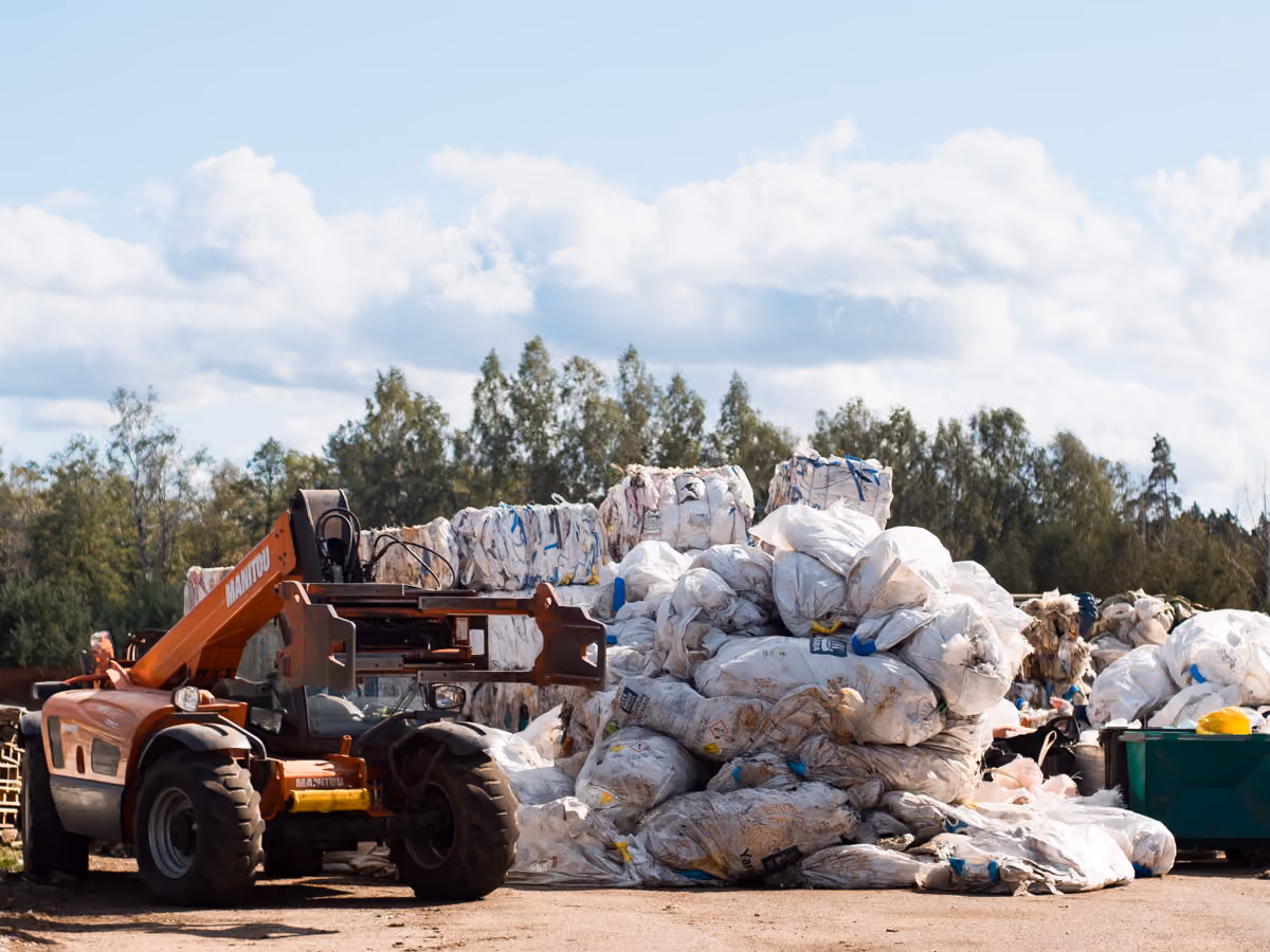Orange forklift near large piles of white wrapped bales outdoors with trees and a cloudy sky behind.