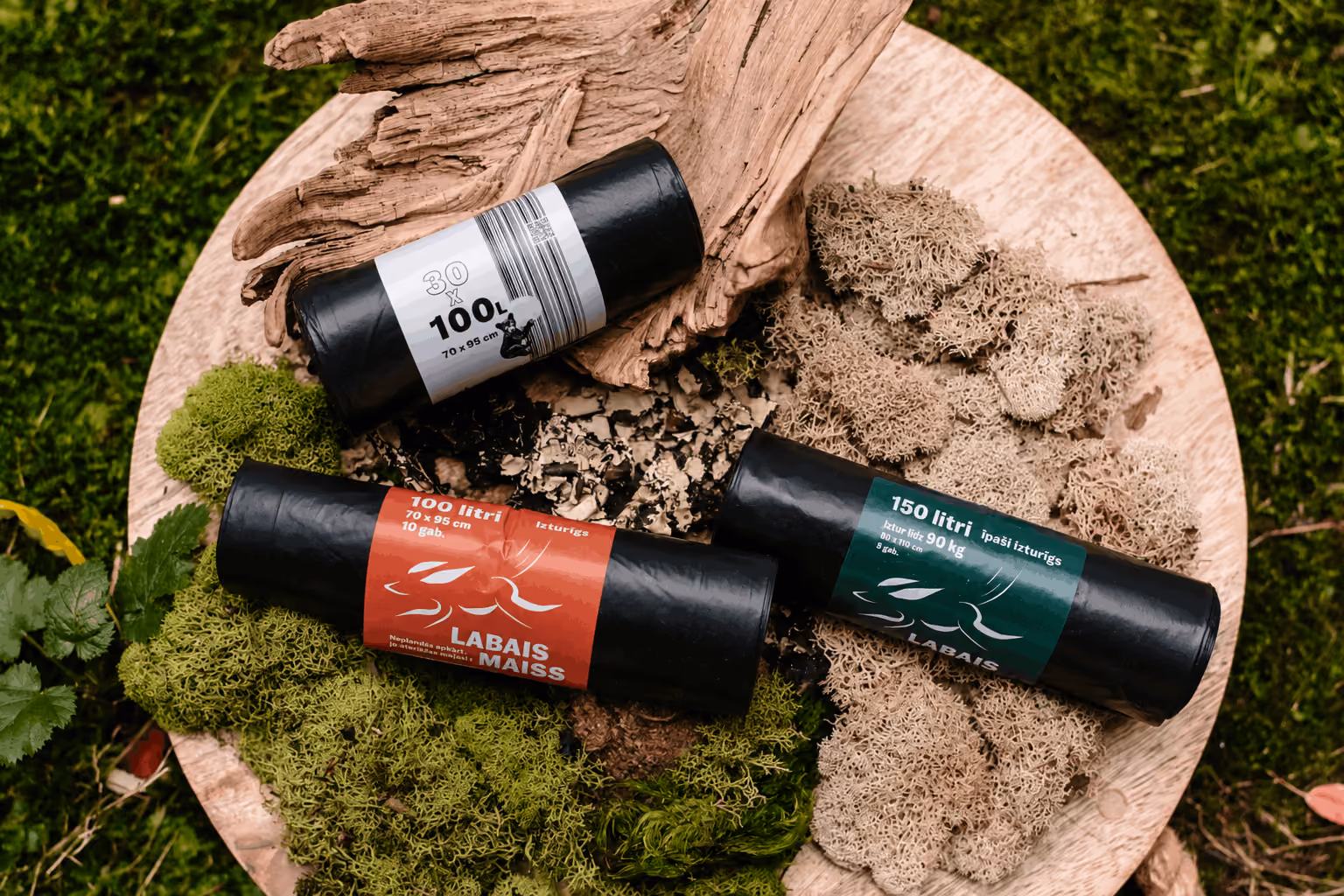 Three rolled black trash bags with different volume labels on a wooden tray with moss and bark.