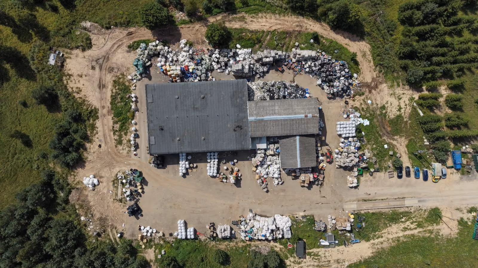 Aerial view of a large warehouse surrounded by piles of wrapped materials and parked vehicles on dirt roads.