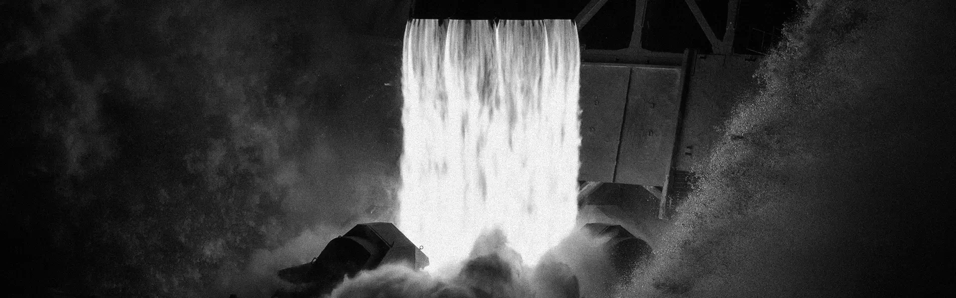 High-contrast black and white image of water forcefully pouring from a sluice gate creating mist and splashes.