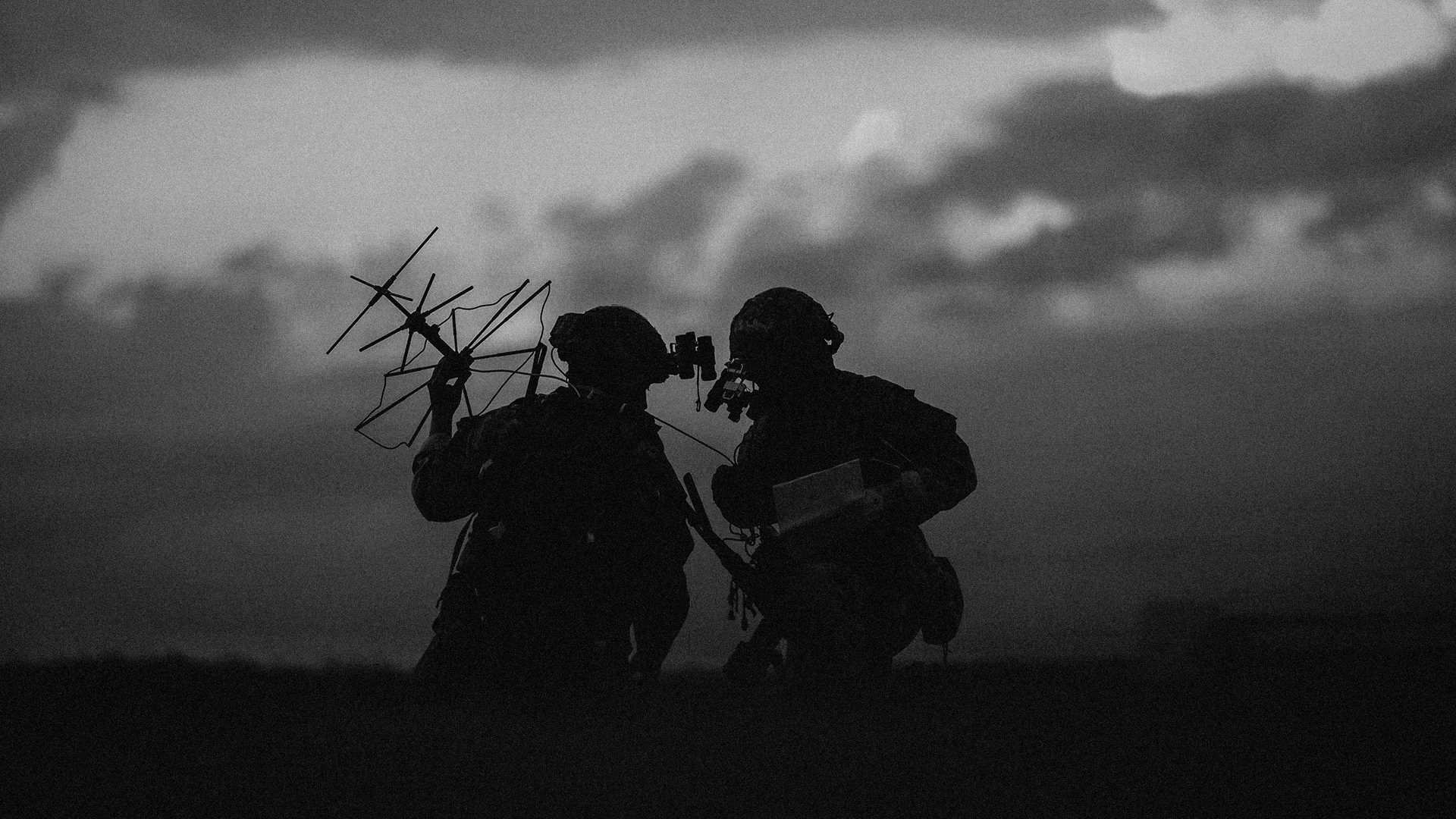 Silhouettes of two soldiers with tactical gear and night vision goggles against a cloudy sky at dusk.