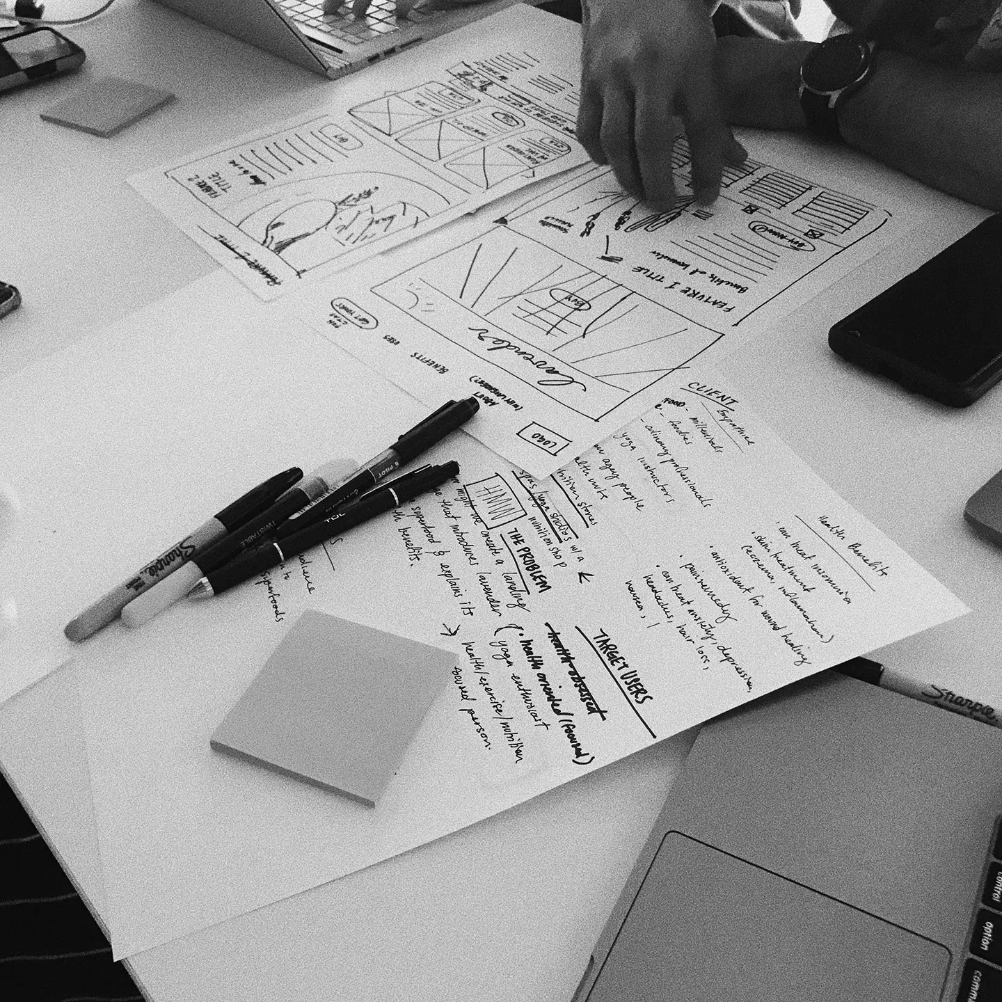 Overhead view of a workspace with printed user interface sketches, handwritten notes, pens, sticky notes, a laptop, and a person's hand pointing at one paper.