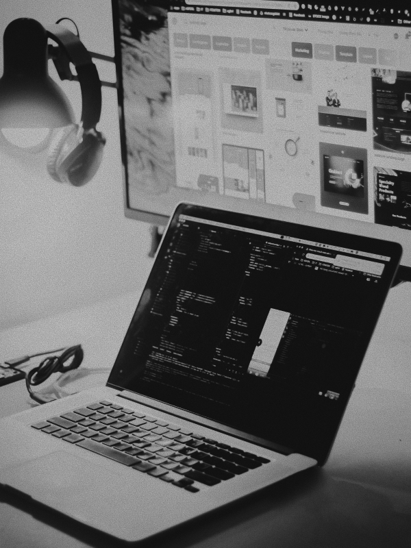 Laptop displaying code on a desk next to a monitor showing a design interface, with headphones hanging on the side.