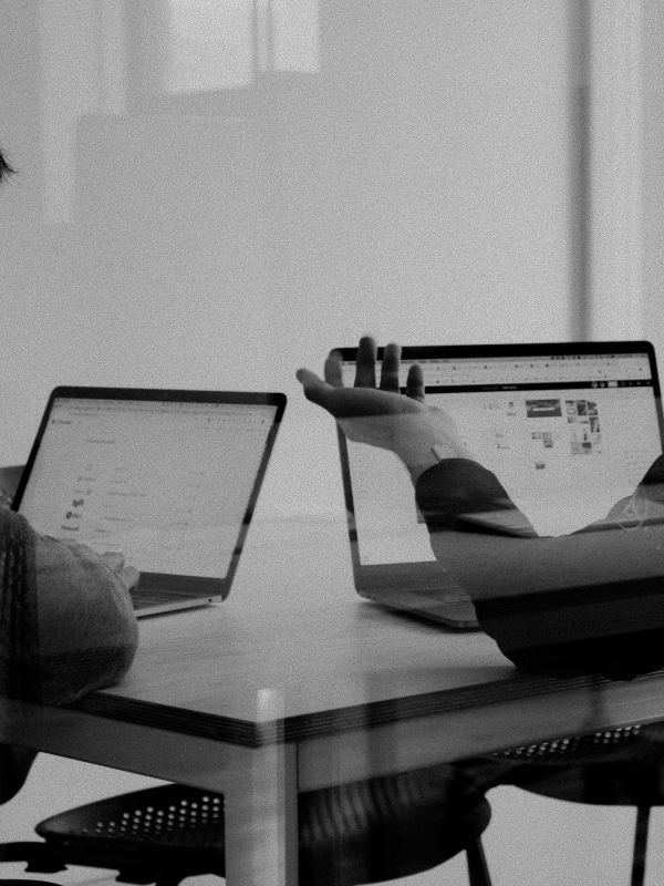 Two people seated at a table working on laptops during a discussion, one person gesturing with an open hand.