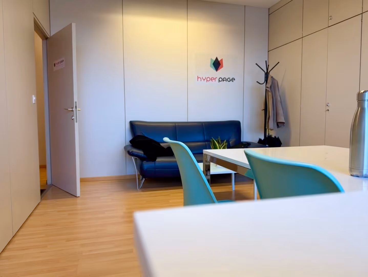 Helles Büro mit Holzboden, blaue Stühle, weißem Tisch, schwarzer Ledercouch und Garderobe mit Jacken.