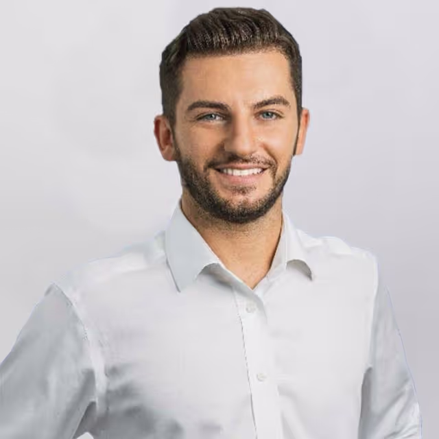 Junger Mann mit kurzen, dunklen Haaren und leichtem Bart, der ein weisses Hemd trägt und lächelt.