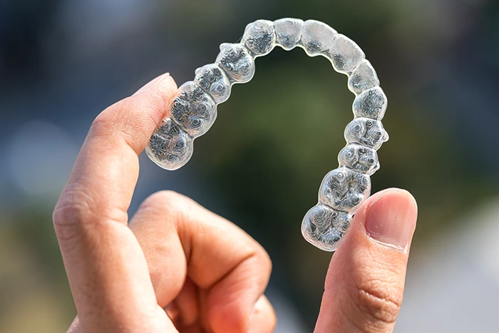 Close-up of a hand holding a transparent dental aligner outdoors against a blurred background.