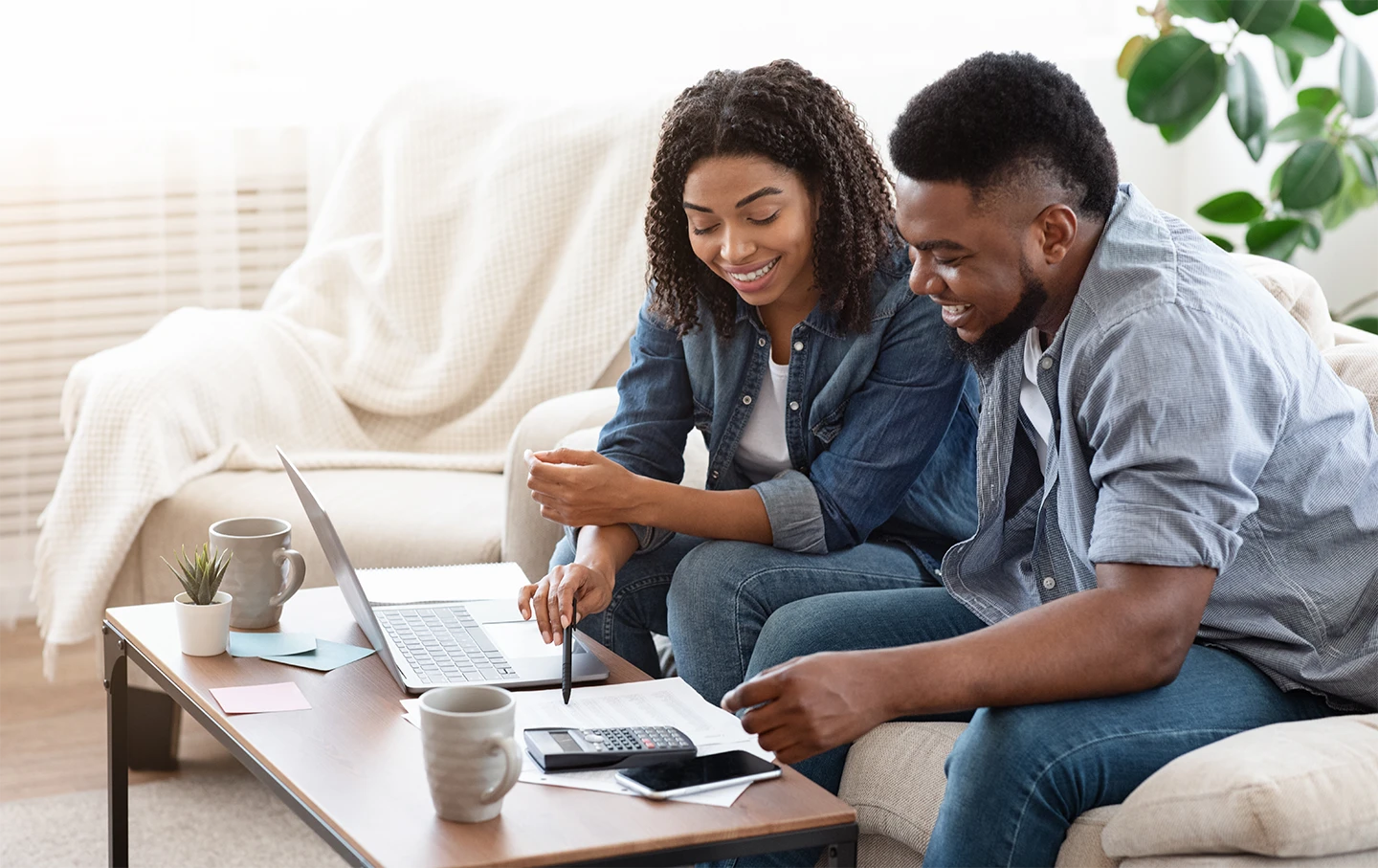 Smiling couple sitting on a couch working on finances with a laptop, papers, calculator, and coffee mugs on a table.