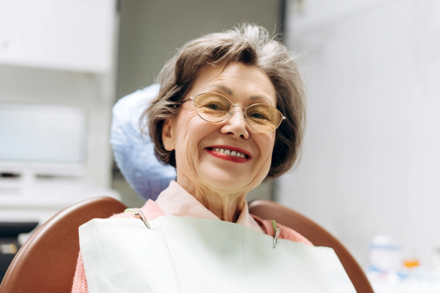 Smiling elderly woman wearing glasses sitting in a dental chair with a dental bib.
