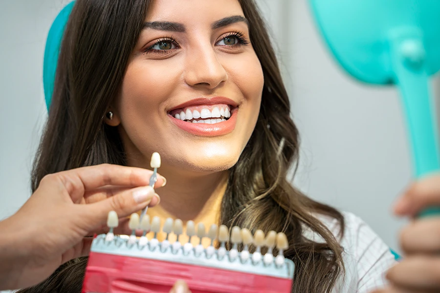 Smiling woman comparing her teeth to a dental shade guide while holding a mirror.