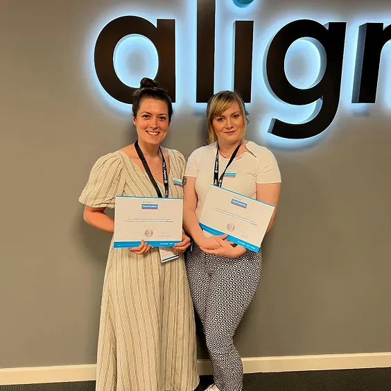 Two women standing and holding certificates in front of a wall with part of the word 'align' displayed.