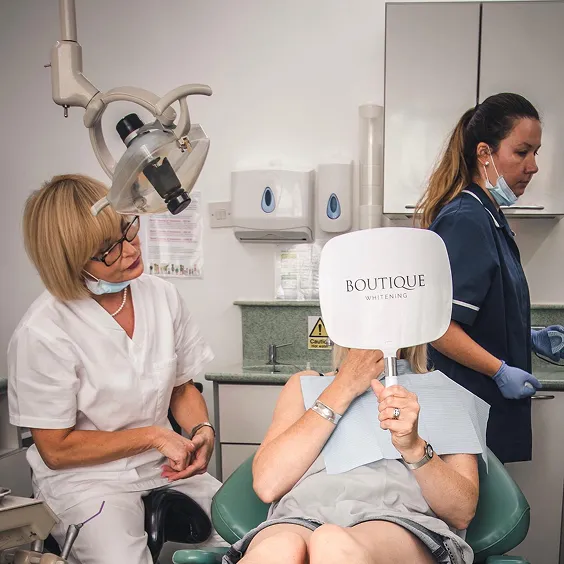 Dentist and assistant attending to a seated patient who holds a Boutique Whitening mirror in a dental clinic.